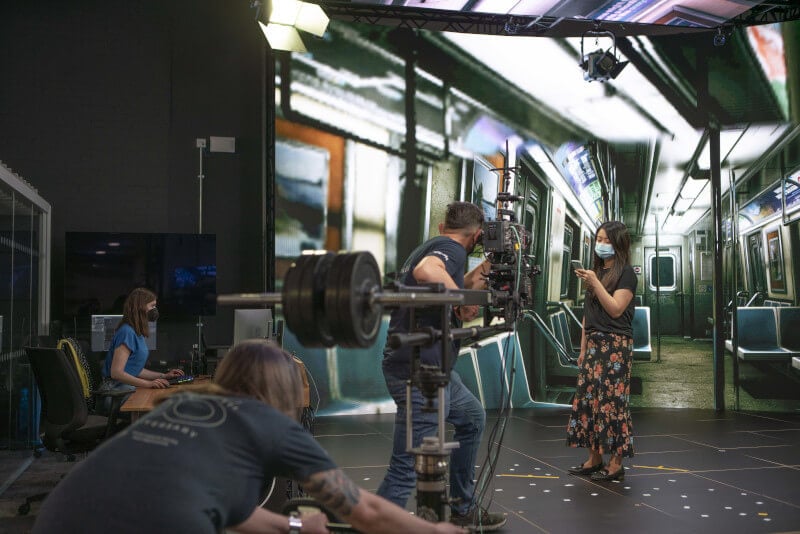 An actress films a scene with a underground train being projected on a screen behind her, being controlled by a technician in the corner